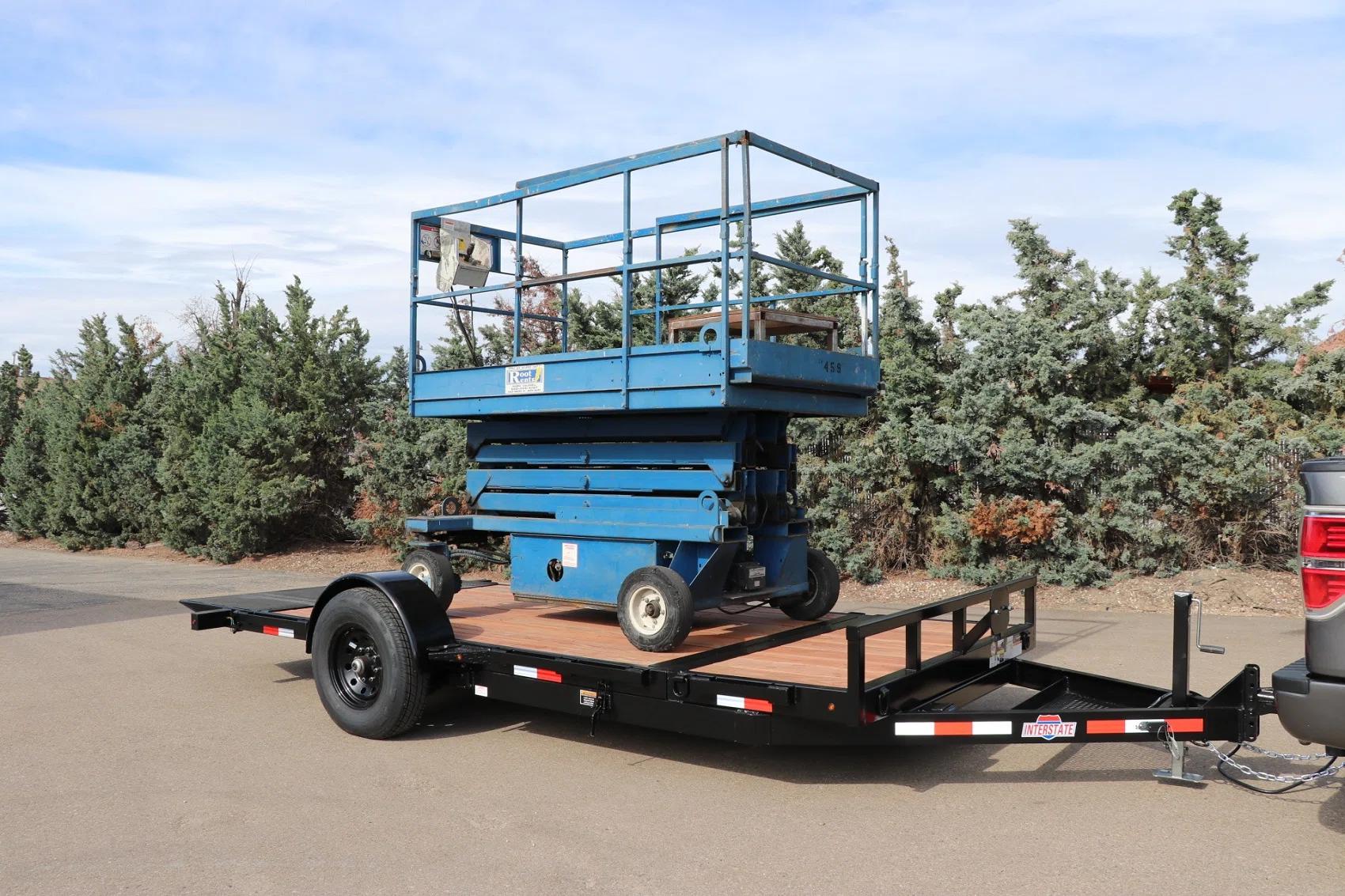 Blue scissor lift loaded onto a black tilt-deck equipment trailer with wood floor, parked at a sunny dealer lot