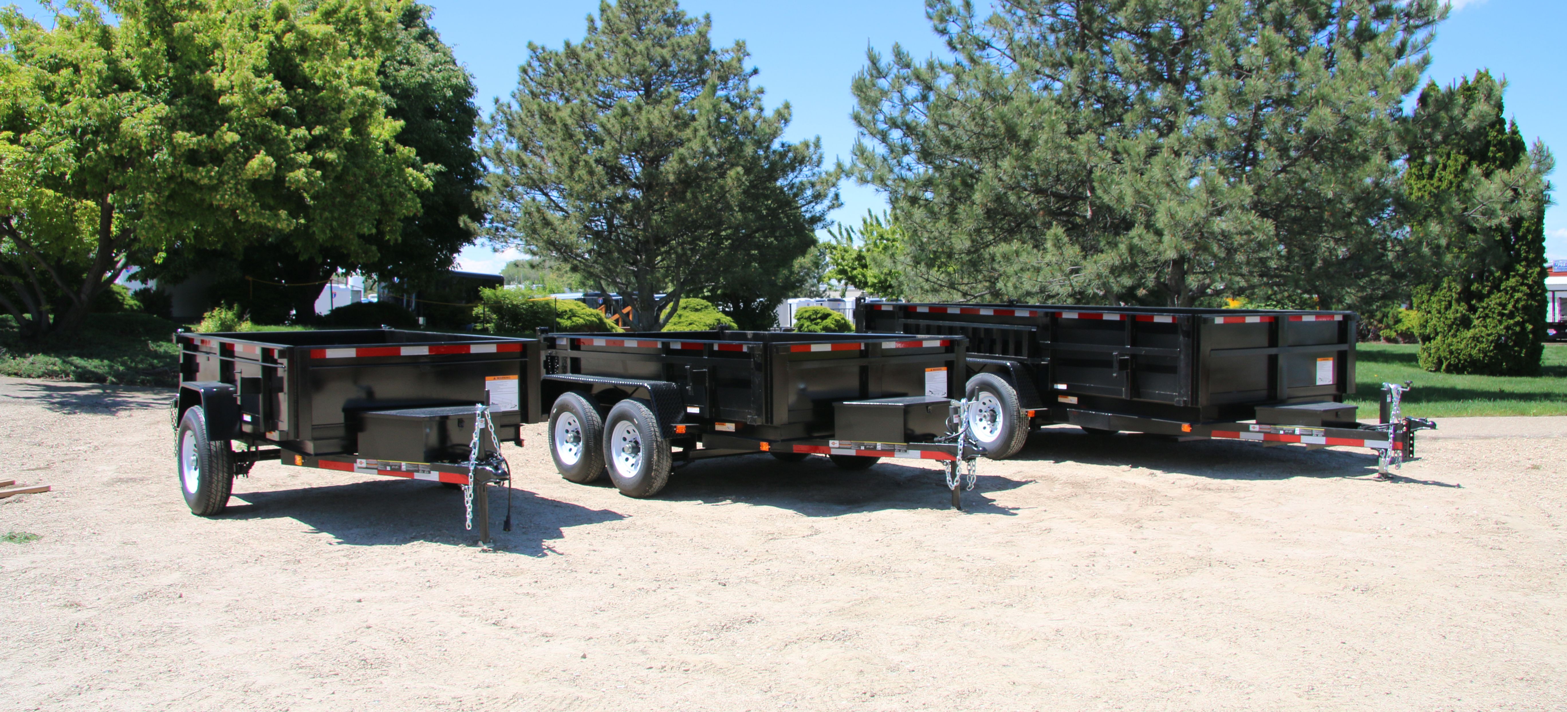 Lineup of a black utility trailer, flatbed deckover, gooseneck dump, and enclosed cargo trailers parked together on a TrailersPlus dealer lot under cloudy skies