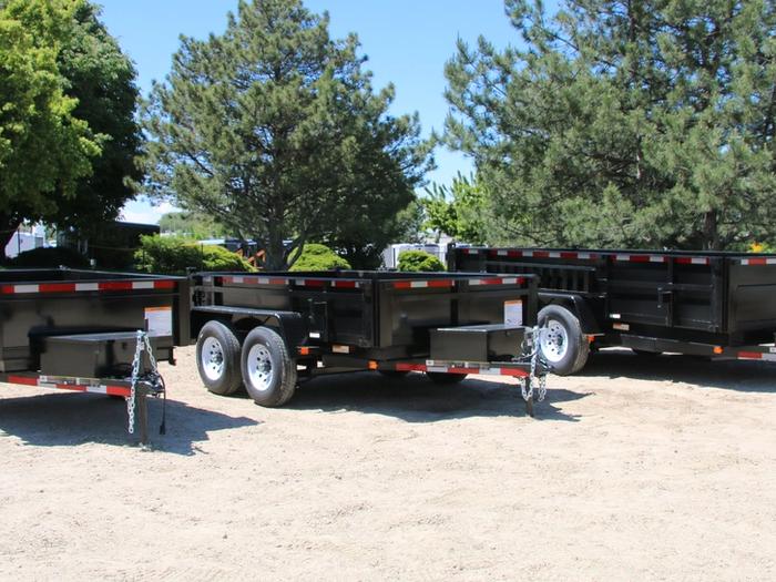 Lineup of a black utility trailer, flatbed deckover, gooseneck dump, and enclosed cargo trailers parked together on a TrailersPlus dealer lot under cloudy skies