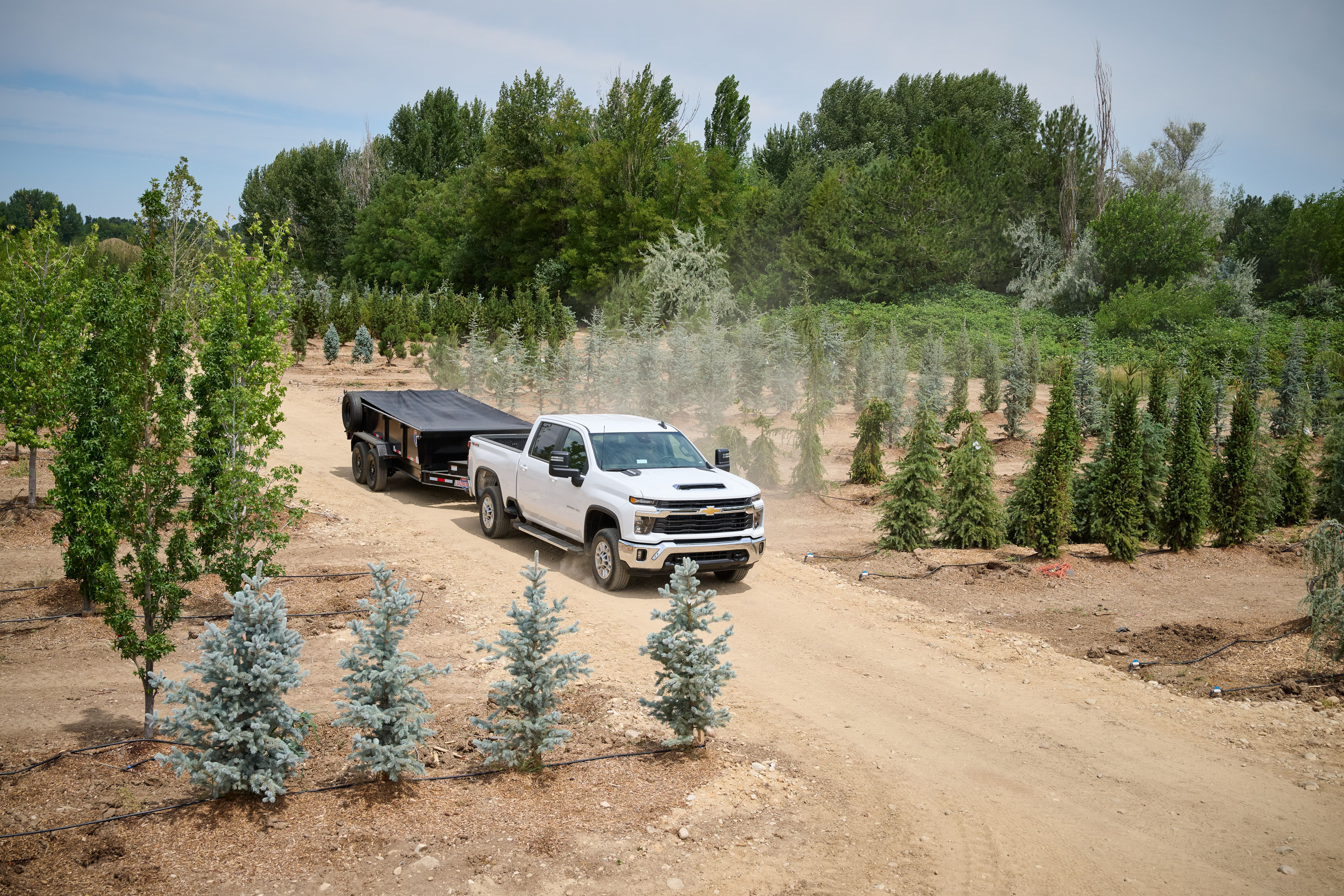 White chevrolet truck driving an enclosed trailer on a dirt road through green trees