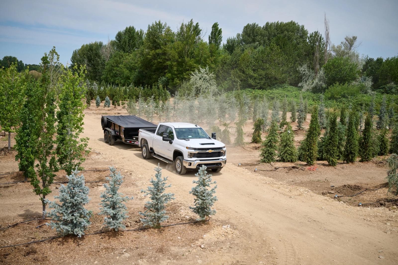 White chevrolet truck driving an enclosed trailer on a dirt road through green trees