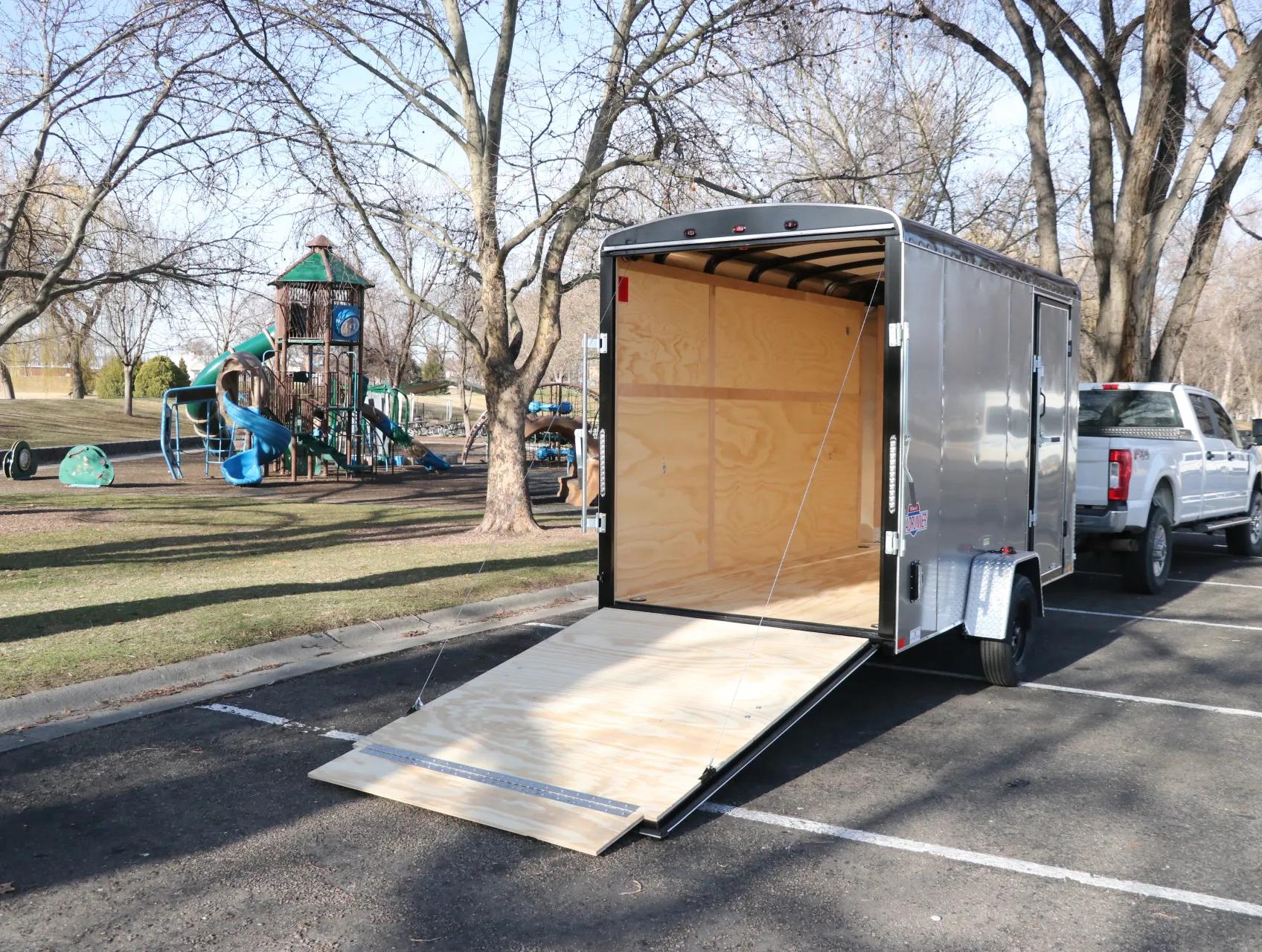Charcoal Interstate enclosed cargo trailer with rear ramp deployed in a paved lot beside a children's playground and bare trees
