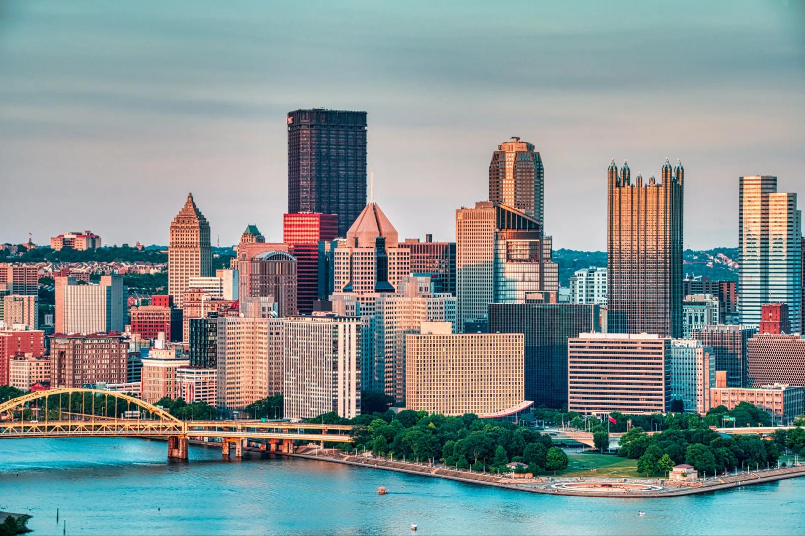 Pittsburgh skyline at golden hour with high-rises along the river and the green Fort Pitt Bridge crossing in the foreground