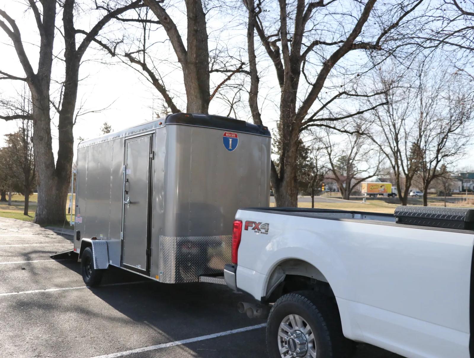 Charcoal Interstate enclosed cargo trailer hitched behind a white pickup truck parked under bare trees in a paved lot