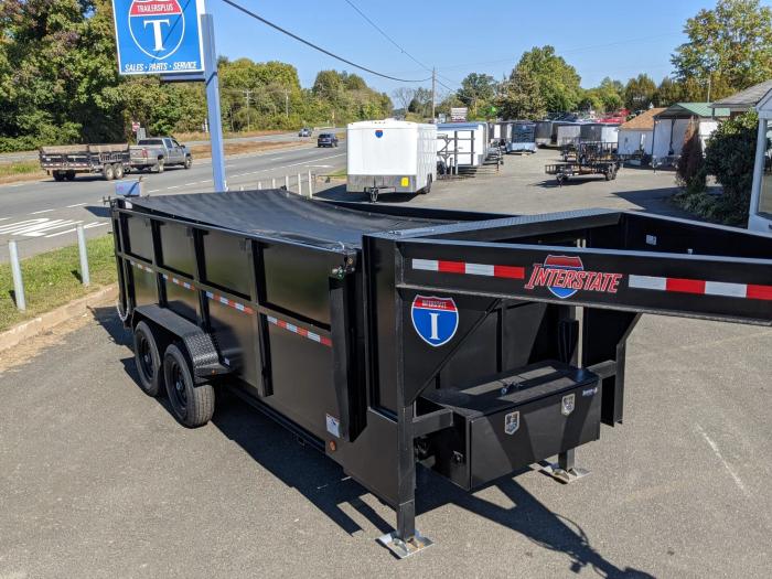 Black tandem-axle dump trailer with a rolled tarp cover installed, parked at a TrailersPlus dealer beside a roadway with other trailers in the background