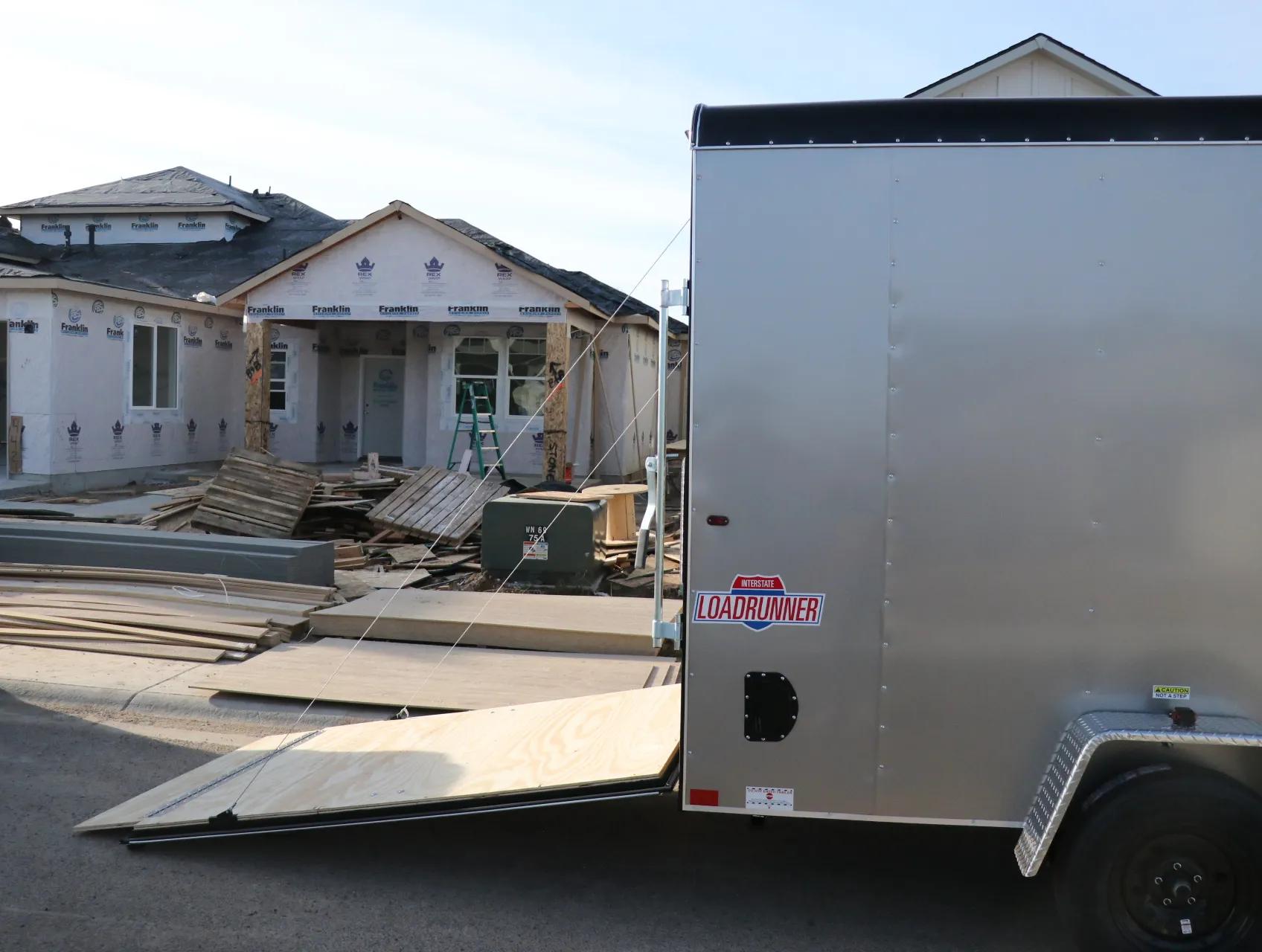 Black single-axle utility trailer with wood deck and rear mesh ramp gate parked on gravel in front of a blue building