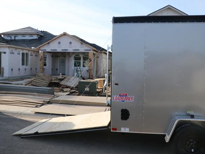 Black single-axle utility trailer with wood deck and rear mesh ramp gate parked on gravel in front of a blue building