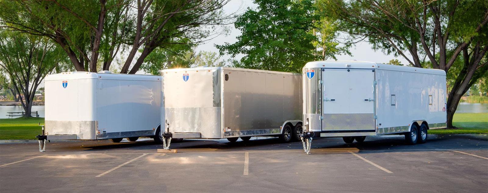 Three white Interstate enclosed cargo trailers parked at angled spaces in a tree-lined paved lot