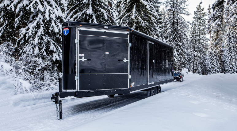 Lineup of a black utility trailer, flatbed deckover, gooseneck dump, and enclosed cargo trailers parked together on a TrailersPlus dealer lot under cloudy skies