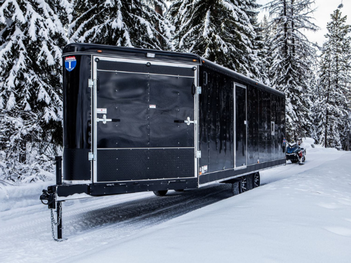 Lineup of a black utility trailer, flatbed deckover, gooseneck dump, and enclosed cargo trailers parked together on a TrailersPlus dealer lot under cloudy skies