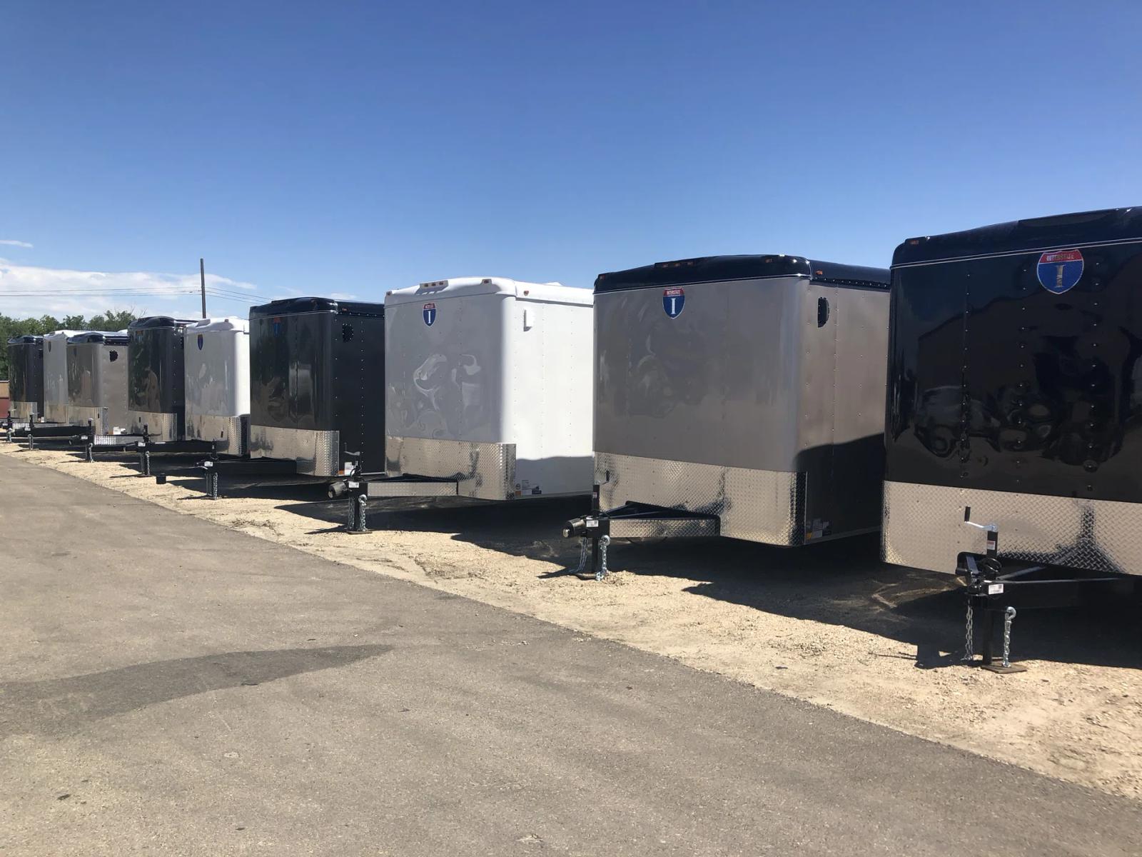 Row of black, silver, and white Interstate enclosed cargo trailers parked along a paved lane at a sunny dealer lot