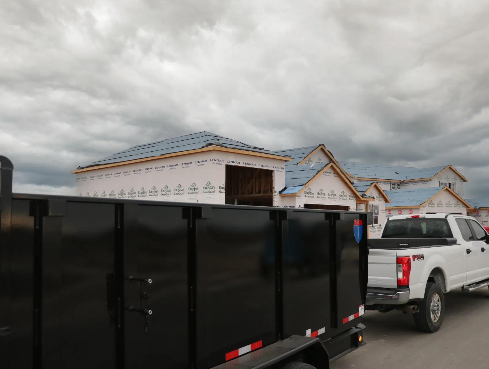 White pickup truck hooked to a tall black dump trailer parked in front of houses under construction on a cloudy day