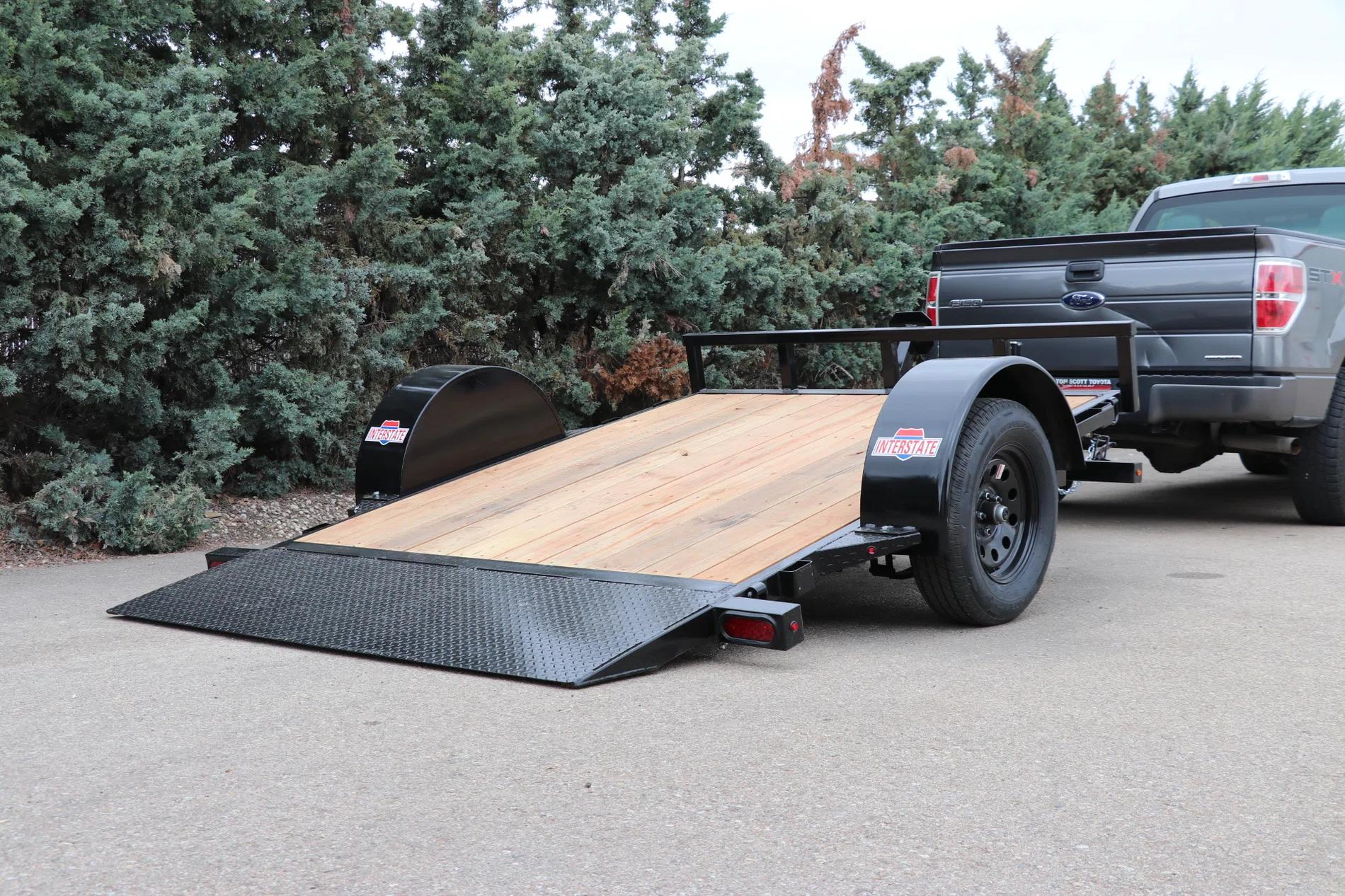 Black single-axle tilt utility trailer with wood deck angled down to ground level, parked behind a black pickup truck