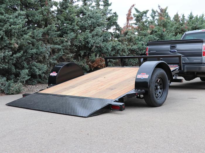 Black single-axle tilt utility trailer with wood deck angled down to ground level, parked behind a black pickup truck