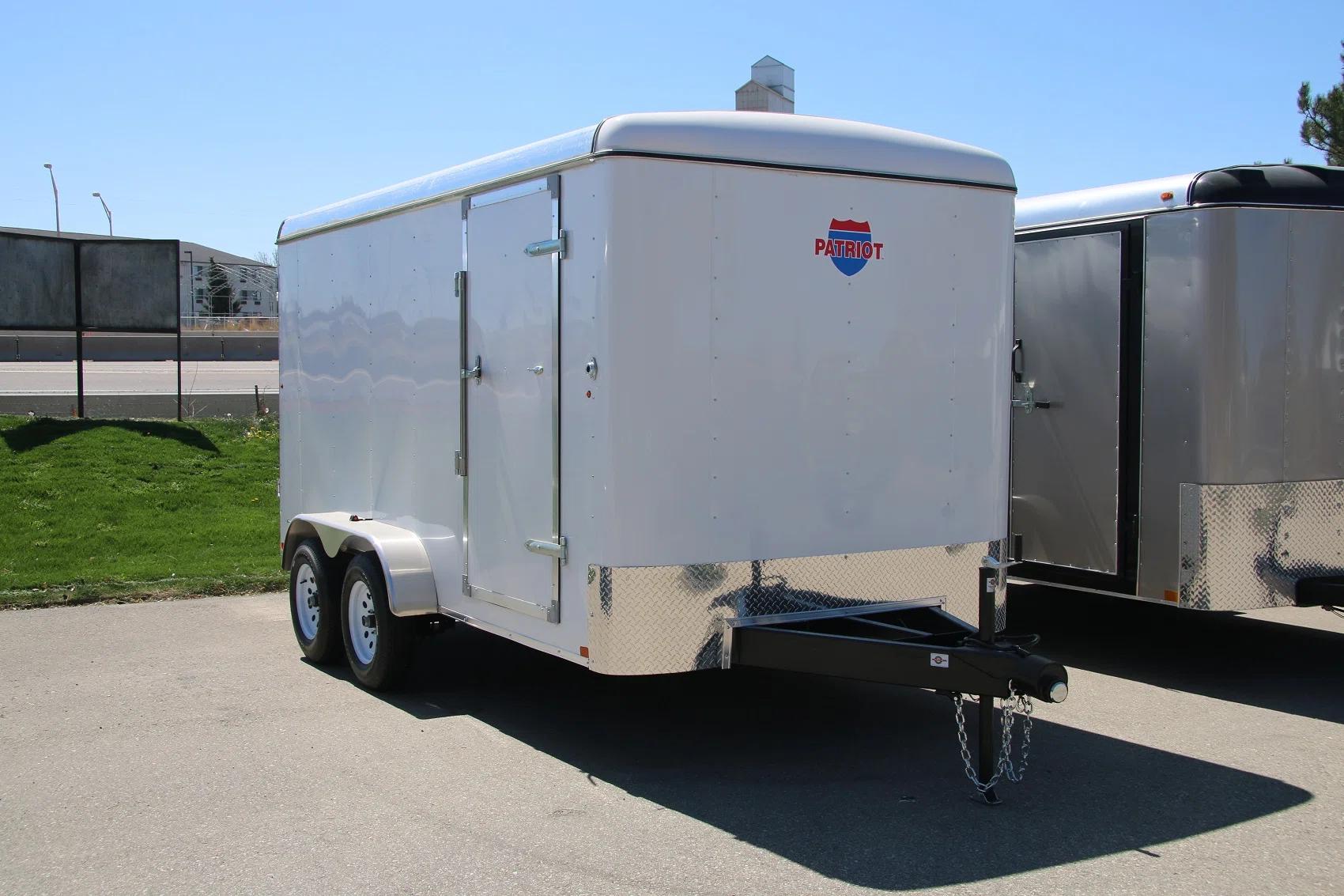 White Patriot-brand enclosed cargo trailer with side door and tandem axles parked on asphalt beside a grassy roadside