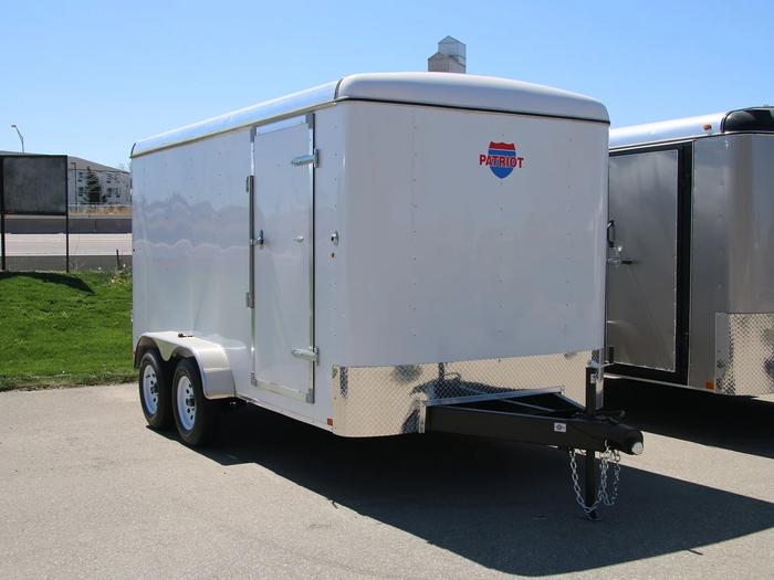 White Patriot-brand enclosed cargo trailer with side door and tandem axles parked on asphalt beside a grassy roadside