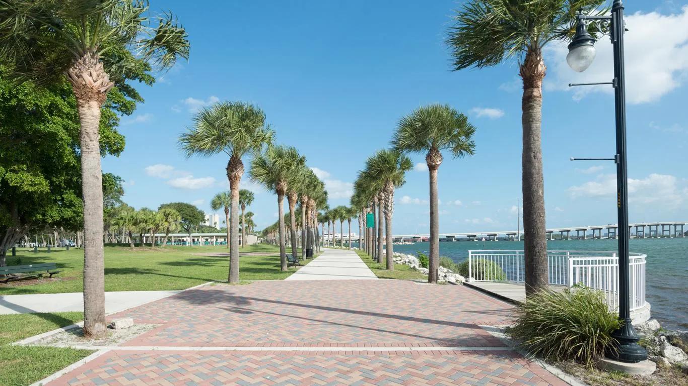 Brick walkway lined with palm trees along a sunny Florida waterfront promenade with a bridge crossing the bay in the distance