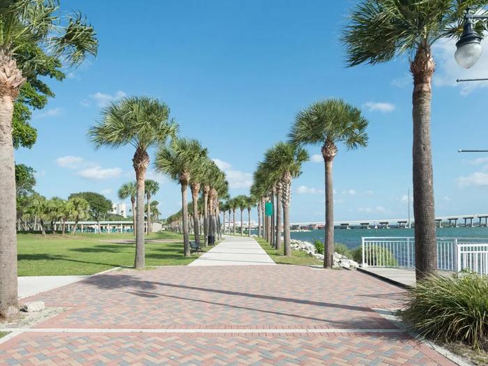 Brick walkway lined with palm trees along a sunny Florida waterfront promenade with a bridge crossing the bay in the distance