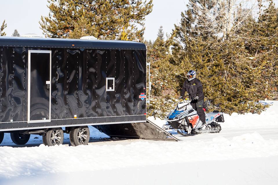 Black enclosed snowmobile trailer with rear ramp deployed in deep snow as a rider stands beside their snowmobile
