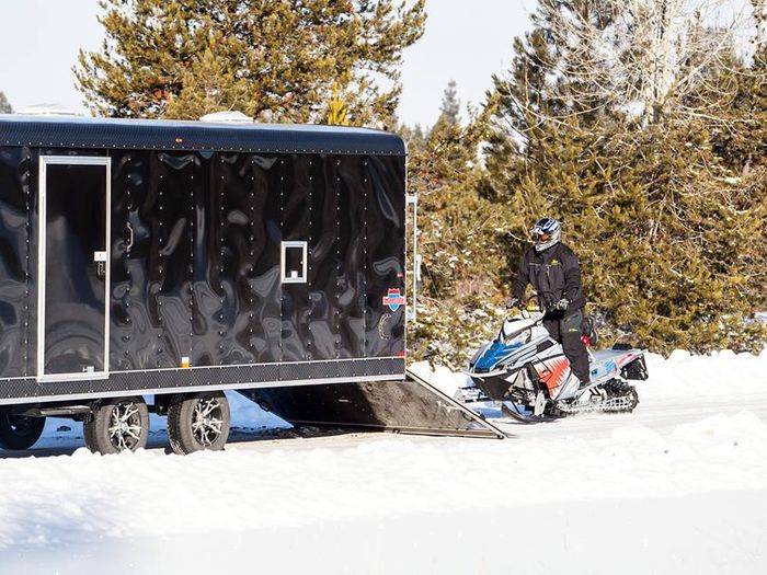 Black enclosed snowmobile trailer with rear ramp deployed in deep snow as a rider stands beside their snowmobile