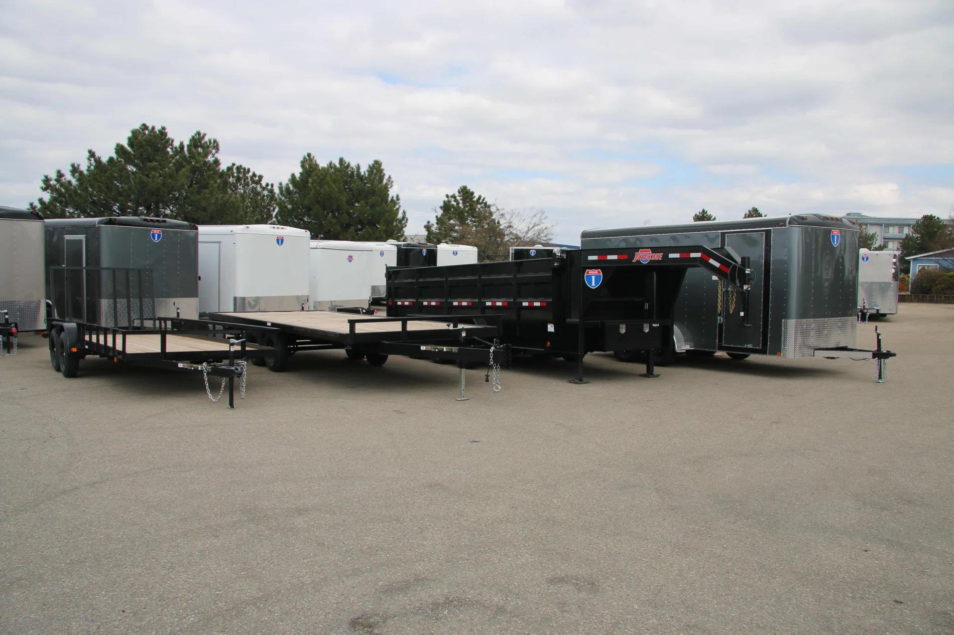 Lineup of a black utility trailer, flatbed deckover, gooseneck dump, and charcoal enclosed cargo trailers parked at a TrailersPlus lot
