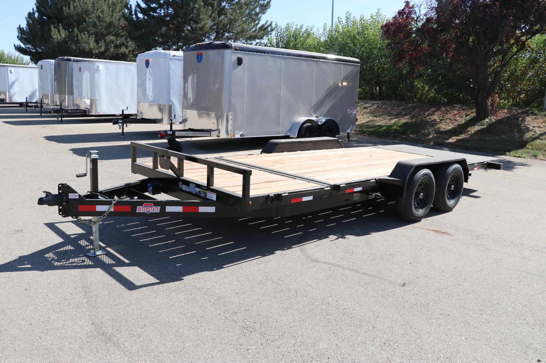 Black tandem-axle flatbed utility trailer with wood deck parked at a TrailersPlus lot with white cargo trailers in the background