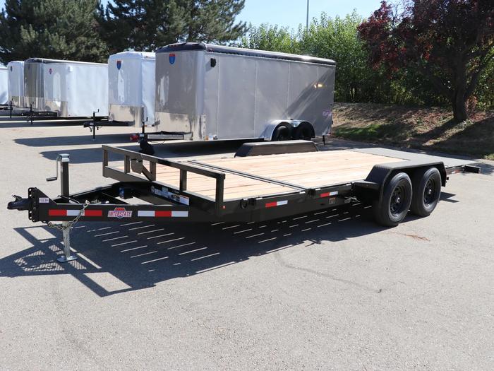 Black tandem-axle flatbed utility trailer with wood deck parked at a TrailersPlus lot with white cargo trailers in the background