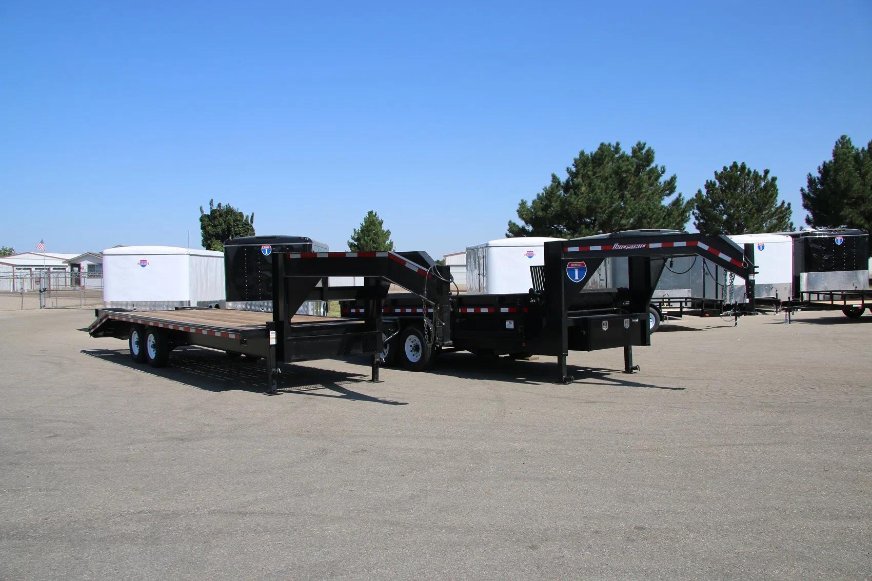 Black tandem-axle dump trailer with the bed tilted up in dump position, parked on gravel at a dealer lot with enclosed cargo trailers in the background