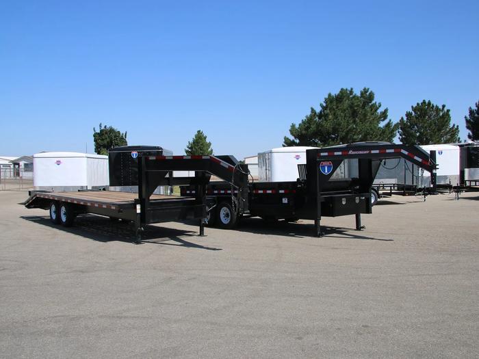 Black tandem-axle dump trailer with the bed tilted up in dump position, parked on gravel at a dealer lot with enclosed cargo trailers in the background
