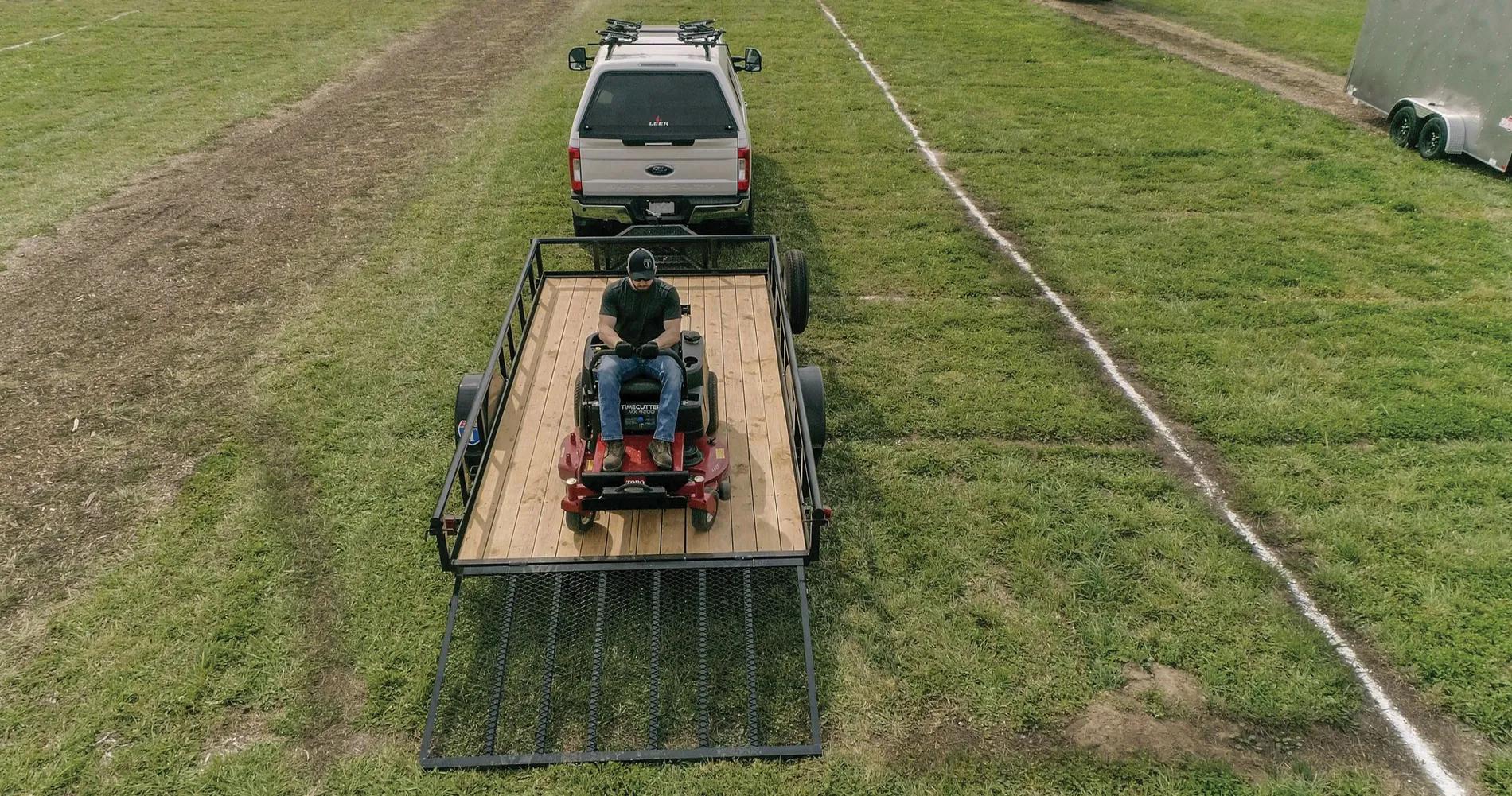 Aerial view of a zero-turn mower loaded on a black utility trailer hitched to a white pickup truck in a green field