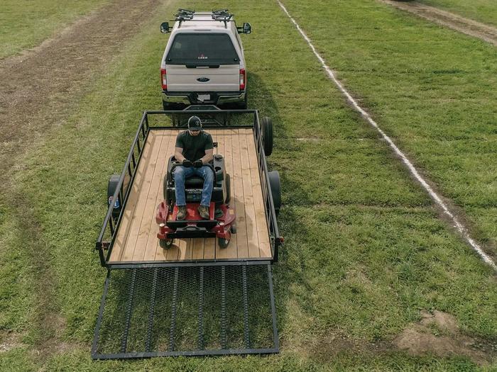 Aerial view of a zero-turn mower loaded on a black utility trailer hitched to a white pickup truck in a green field