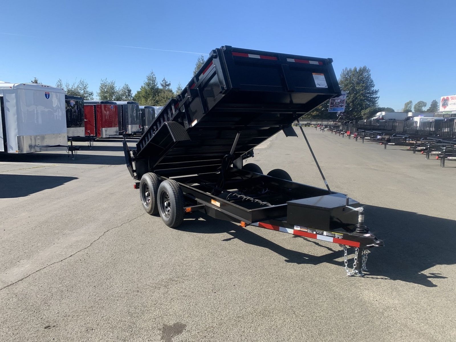 Black tandem-axle dump trailer with its bed raised at full tilt, parked on a TrailersPlus dealer lot with rows of trailers behind it