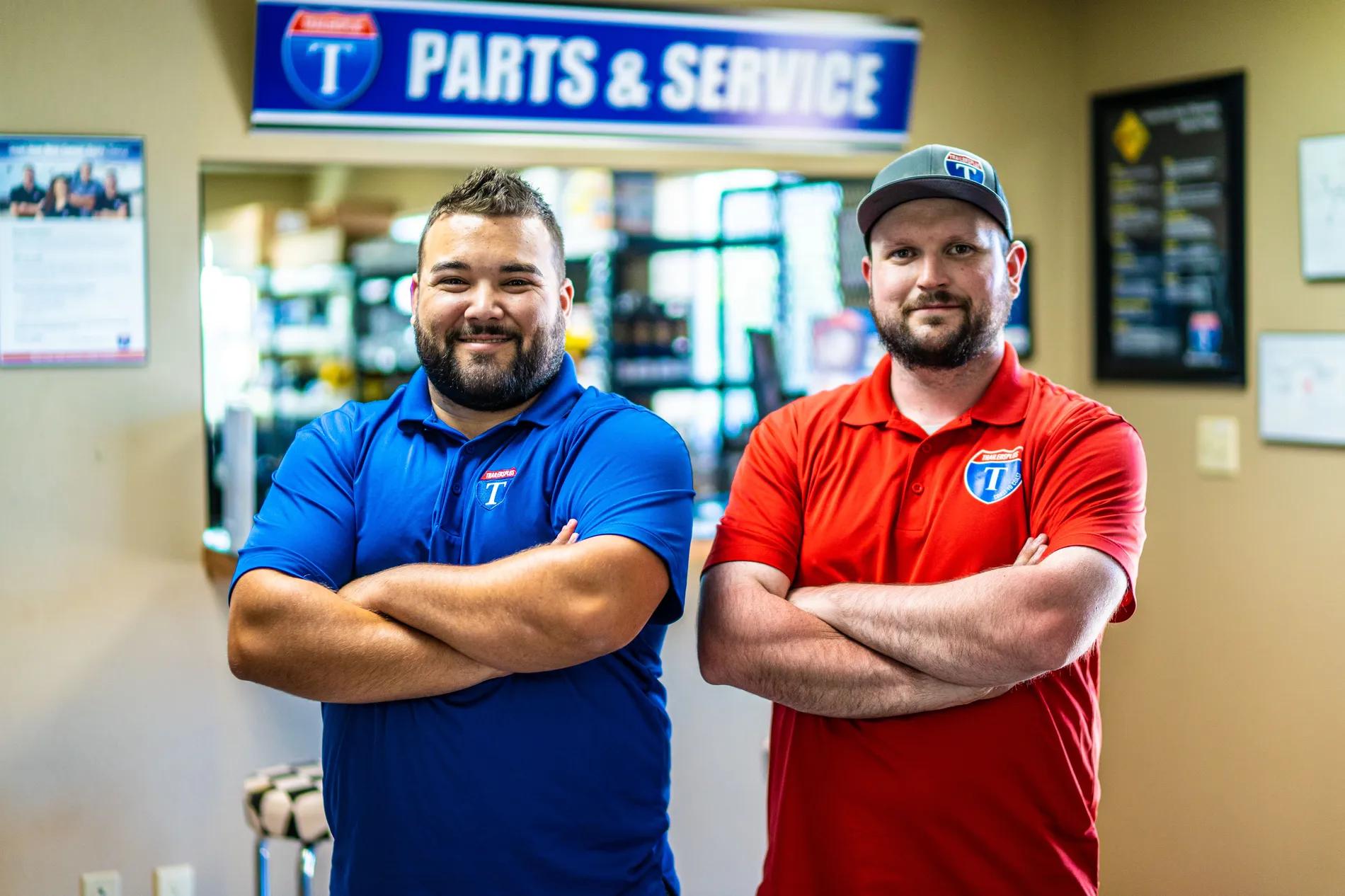 Two smiling TrailersPlus parts and service staff in branded polos standing with arms crossed under the dealership Parts and Service sign