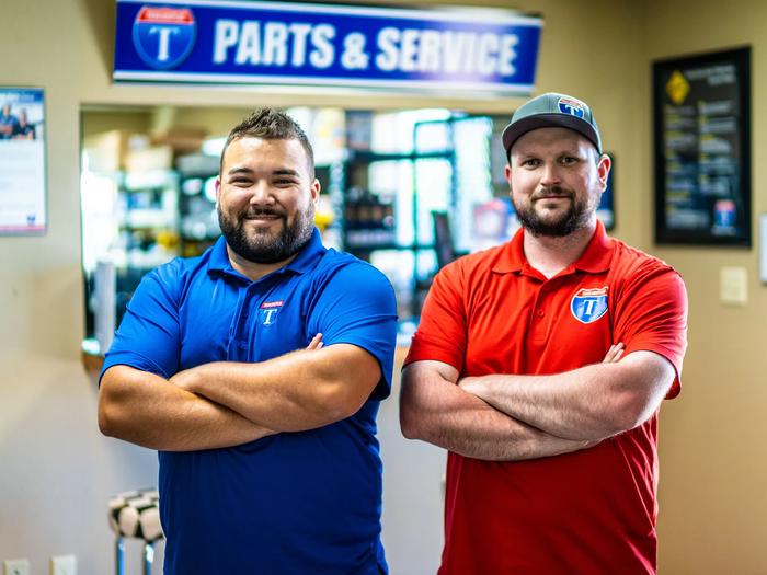 Two smiling TrailersPlus parts and service staff in branded polos standing with arms crossed under the dealership Parts and Service sign