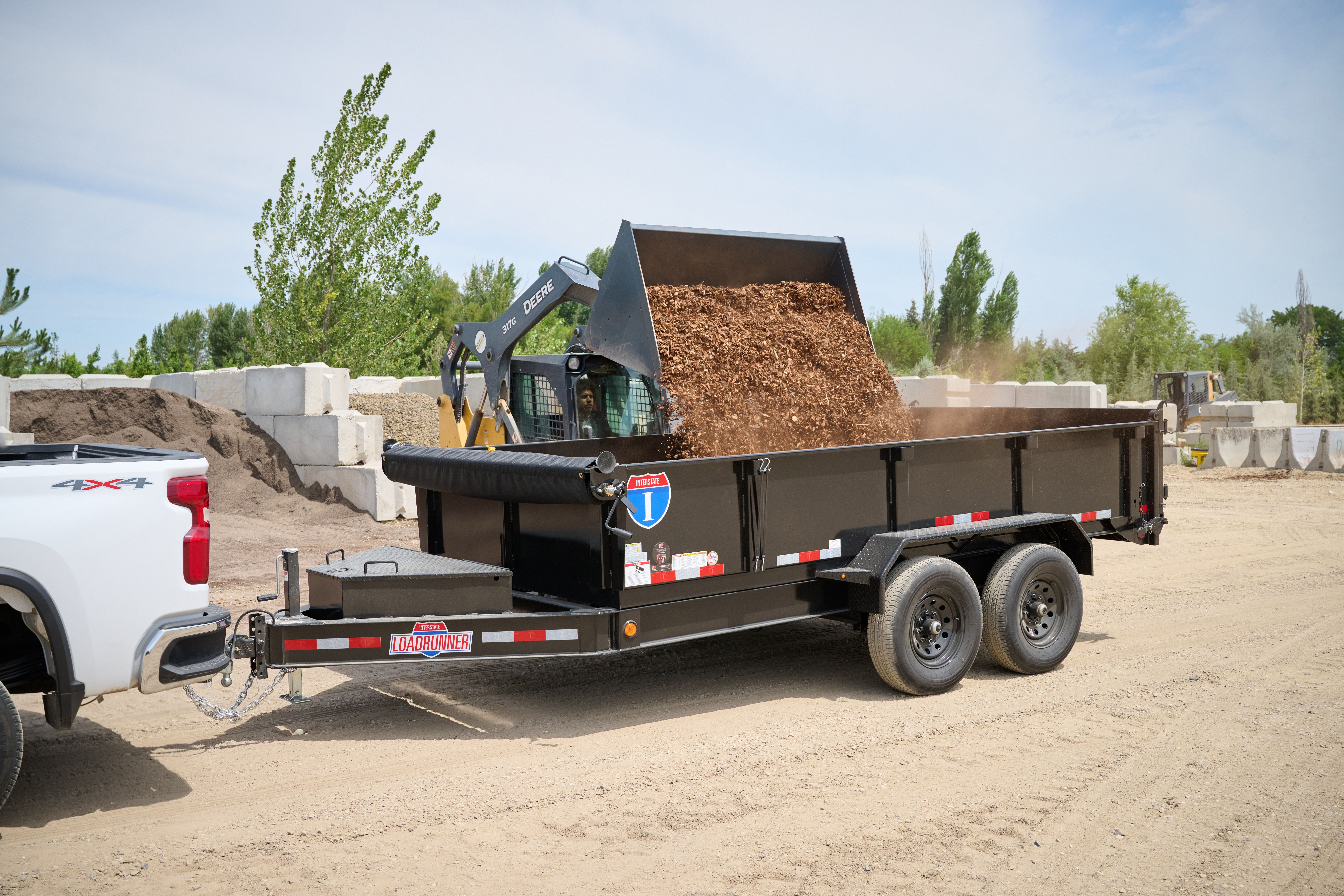 Black dump trailer attached to a white truck, with a Deere skidsteer loading mulch into the dump trailer