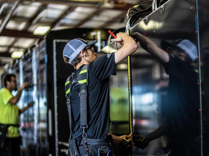 TrailersPlus technician in a hard hat using a tape measure on a black trailer panel inside the dealership service bay