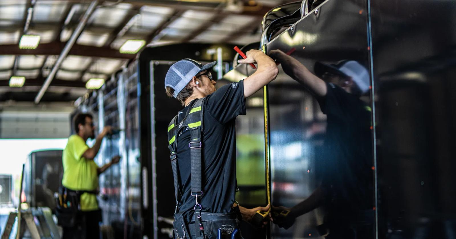 TrailersPlus technician in a hard hat using a tape measure on a black trailer panel inside the dealership service bay
