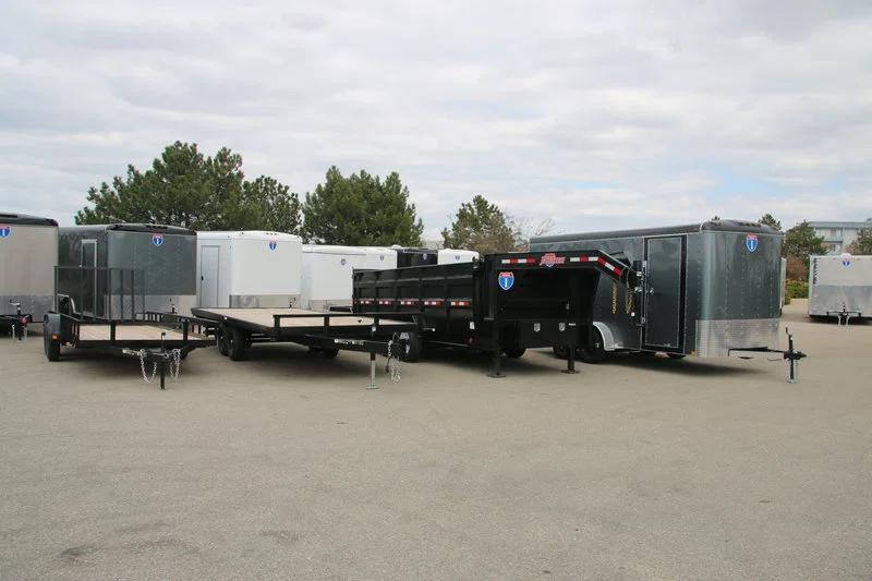 Lineup of a black utility trailer, flatbed deckover, gooseneck dump, and gray enclosed cargo trailers parked together at a TrailersPlus dealer lot