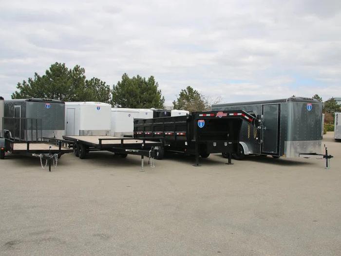 Lineup of a black utility trailer, flatbed deckover, gooseneck dump, and gray enclosed cargo trailers parked together at a TrailersPlus dealer lot