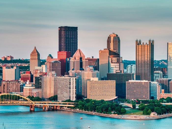 Pittsburgh skyline at golden hour with high-rises along the river and the green Fort Pitt Bridge crossing in the foreground