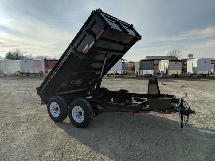 Lineup of a black utility trailer, flatbed deckover, gooseneck dump, and enclosed cargo trailers parked together on a TrailersPlus dealer lot under cloudy skies