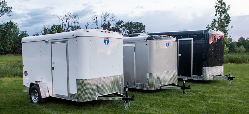 Three white Interstate enclosed cargo trailers parked at angled spaces in a tree-lined paved lot