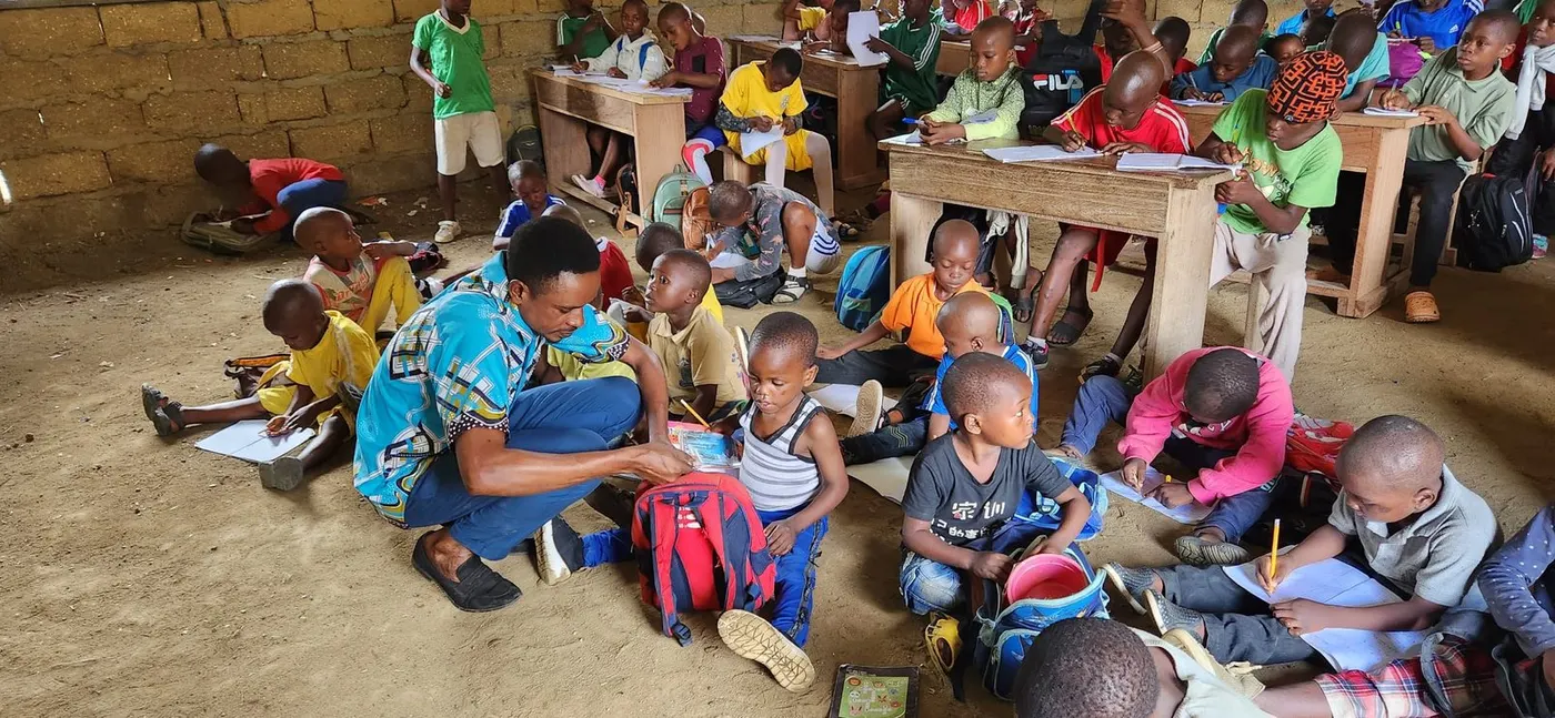 Before the rebuild: pupils writing on the earth floor of a crowded classroom while a teacher helps