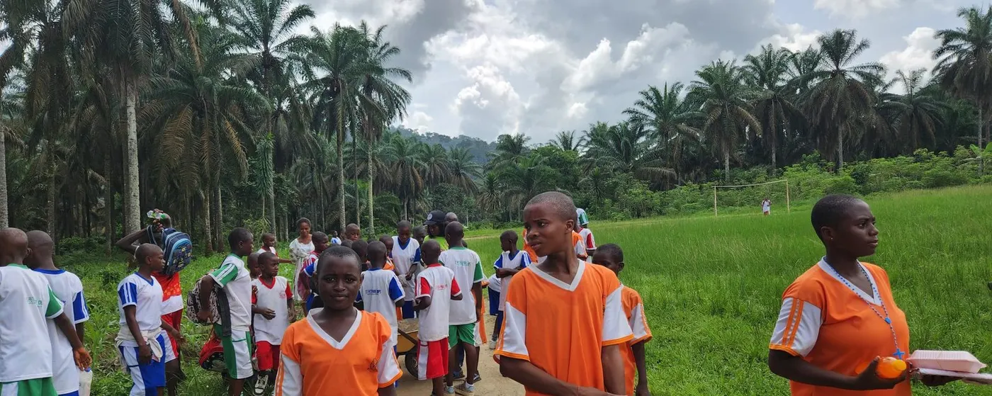 Pupils in orange and white sports kit on the green school field, oil palms behind