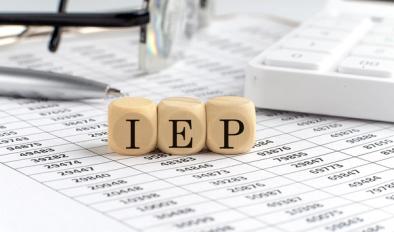 Close-up of wooden blocks spelling IEP, placed on top of a spreadsheet with a pen, calculator, and eyeglasses, representing Individualized Education Program planning and documentation.