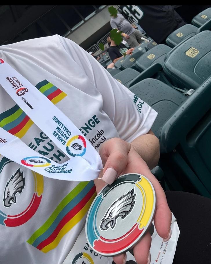 Hand holding Eagles Autism Challenge finisher medal featuring Philadelphia Eagles logo with colorful rainbow design, alongside white Eagles Autism Challenge t-shirt with Lincoln Financial sponsor branding, taken in stadium seating at Lincoln Financial Field
