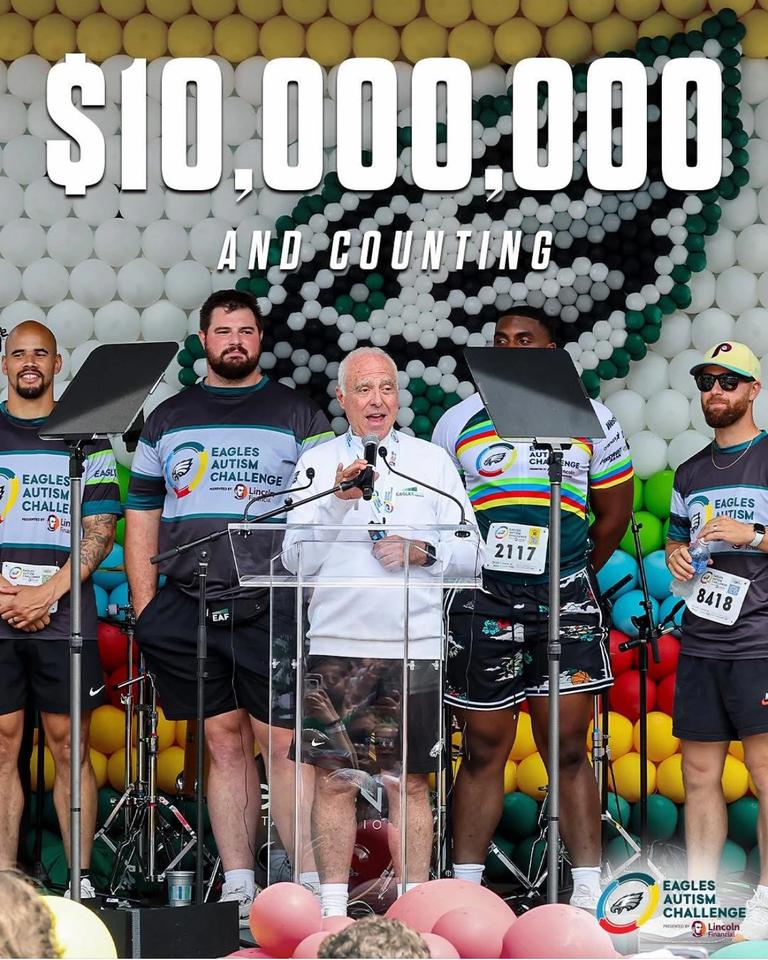 Philadelphia Eagles owner Jeffrey Lurie speaking at podium during Eagles Autism Challenge event, surrounded by participants and athletes wearing Eagles Autism Challenge jerseys, with '$10,000,000 and counting' displayed on balloon backdrop celebrating fundraising milestone.