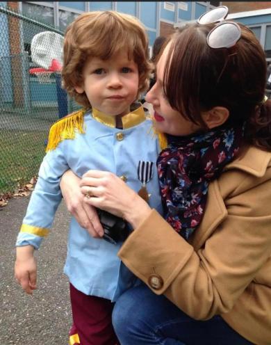 Tracie Smith kneeling beside her young son Halen, who is dressed in a prince costume with golden shoulder tassels, while outdoors near a schoolyard.