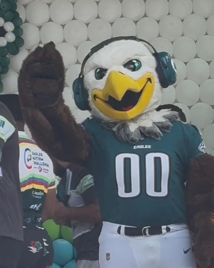 Philadelphia Eagles mascot Swoop wearing headphones and waving while wearing Eagles jersey number 00, surrounded by Eagles Autism Challenge participants and colorful balloon decorations at Lincoln Financial Field