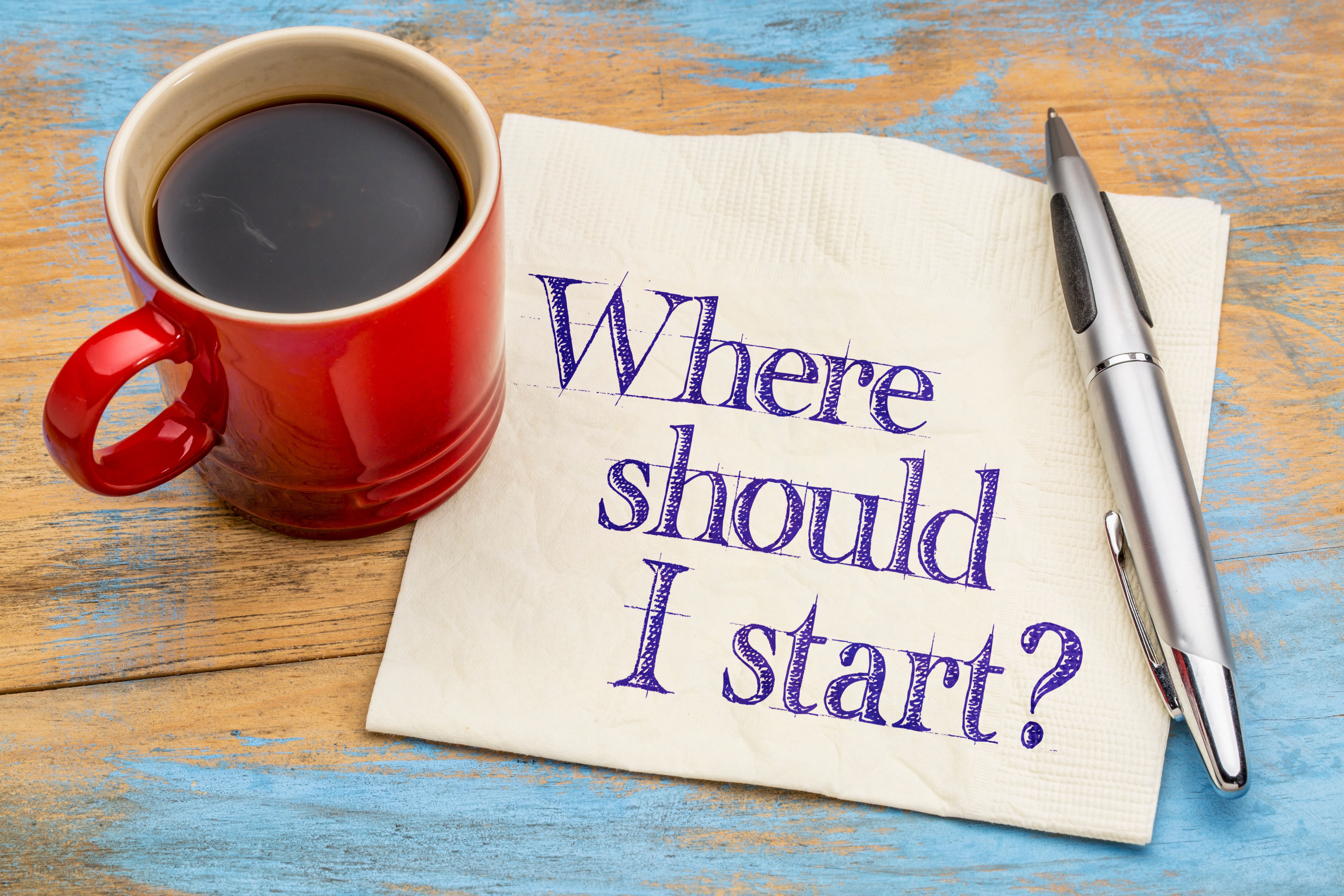 Red coffee cup next to a silver pen and a napkin with the handwritten text 'Where should I start?' placed on a rustic wooden table with blue accents